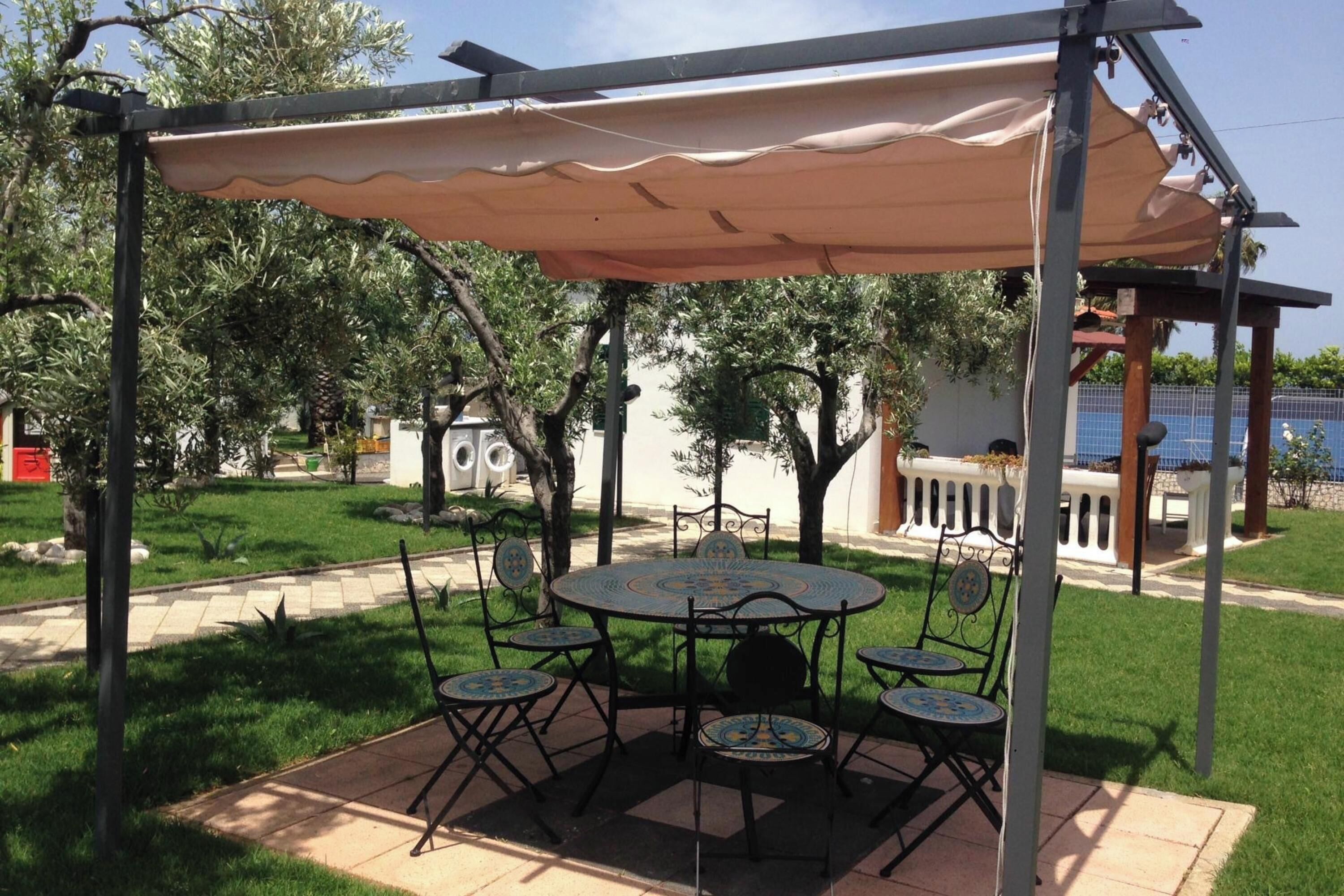 Une terrasse extérieure couverte, avec une table ronde et des chaises, entourée de verdure et d’arbres.