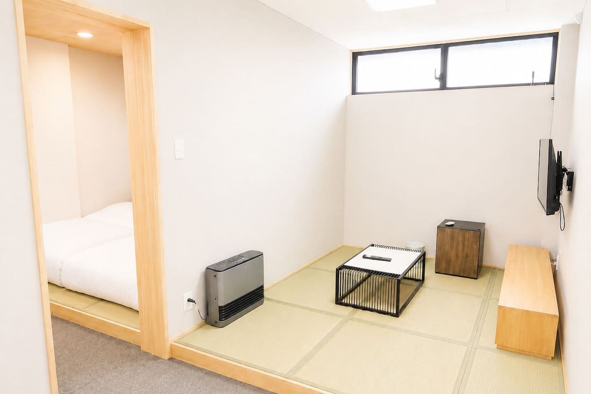 A compact room with a bed, a heater, a small table with a bowl, a wooden cabinet, and a TV mounted on the wall.