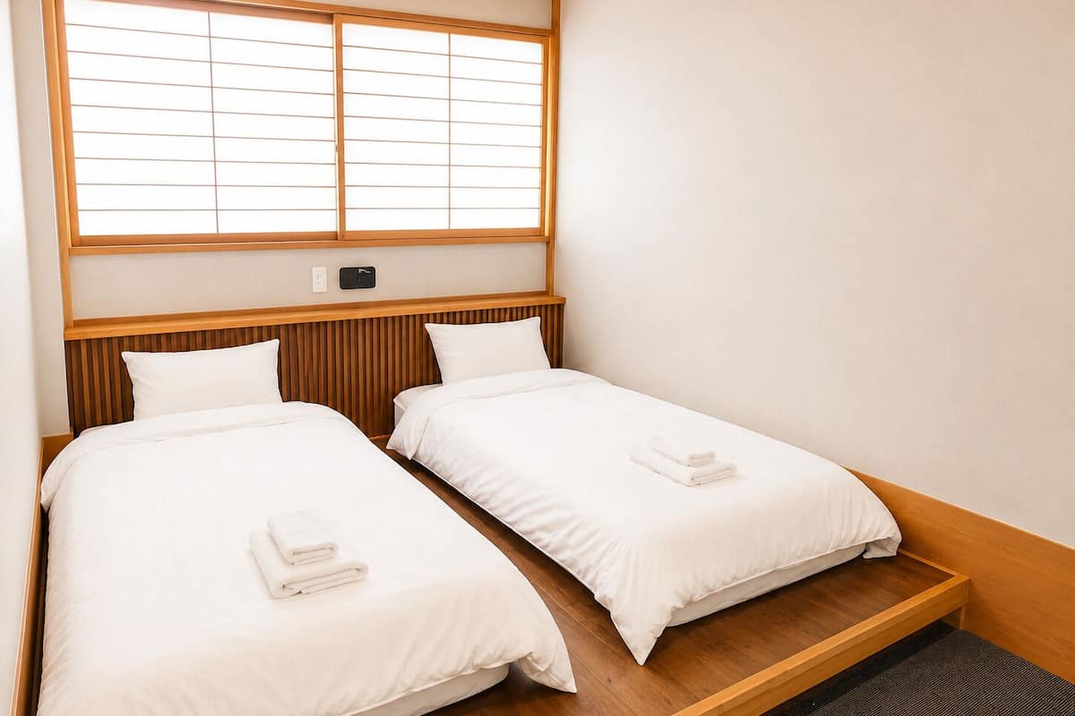 A hotel room with two single beds, wooden headboards, a window with blinds, and a small bedside table.