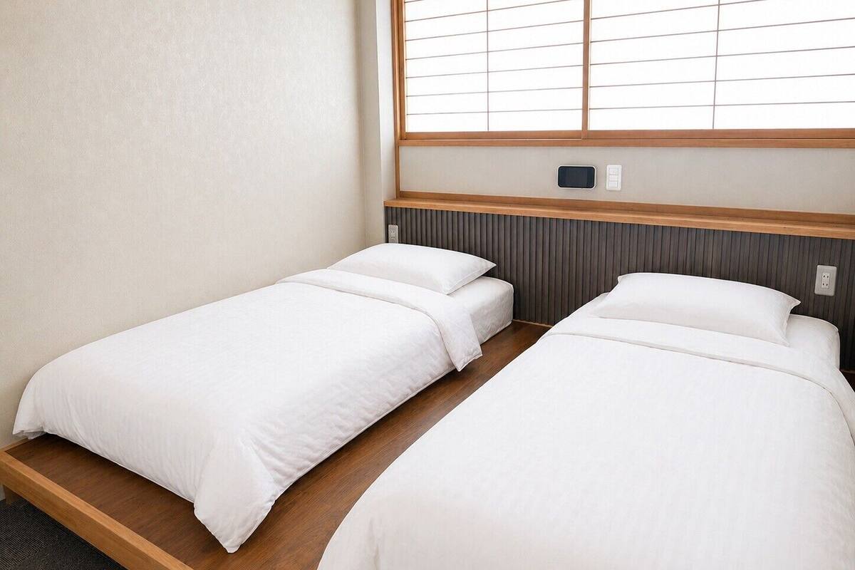 A room with two single beds, a wooden floor, a window with blinds, and a wall-mounted thermostat.