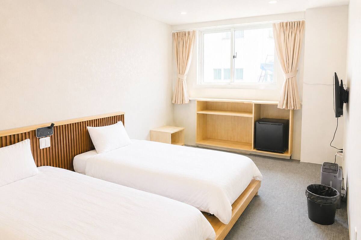 A hotel room with two beds, a TV, a desk, a small storage unit, and a window with curtains.