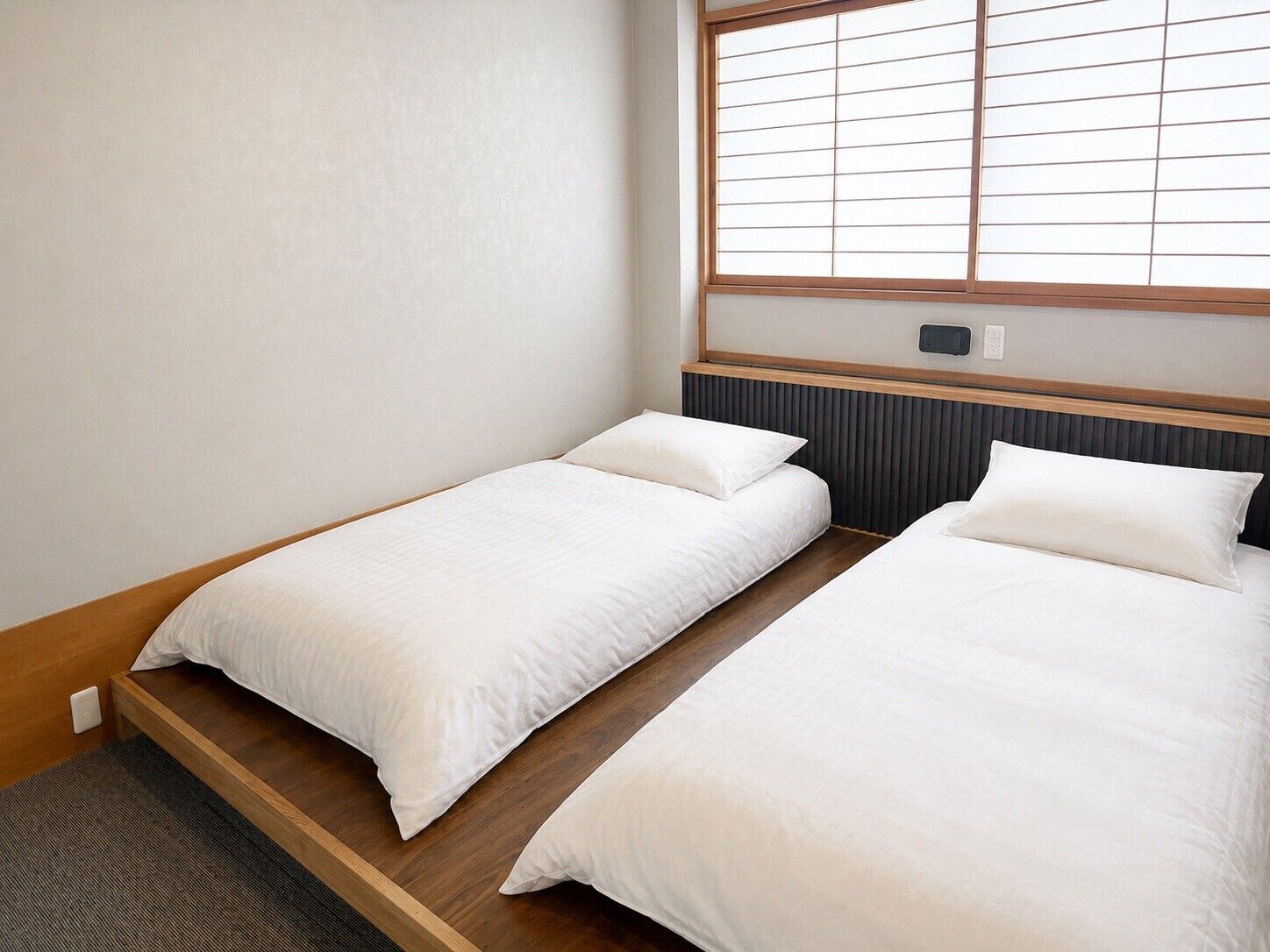 A room with two single beds, a window with horizontal blinds, a wooden headboard, and a small bedside table.