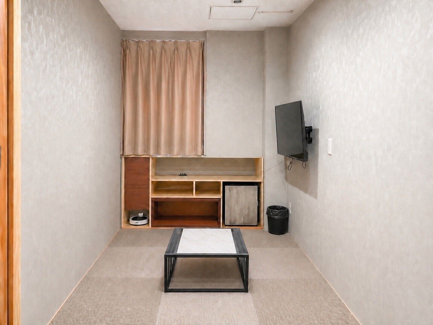 A small, well-lit room with a bed, a TV mounted on the wall, a wooden cabinet, and a small table.
