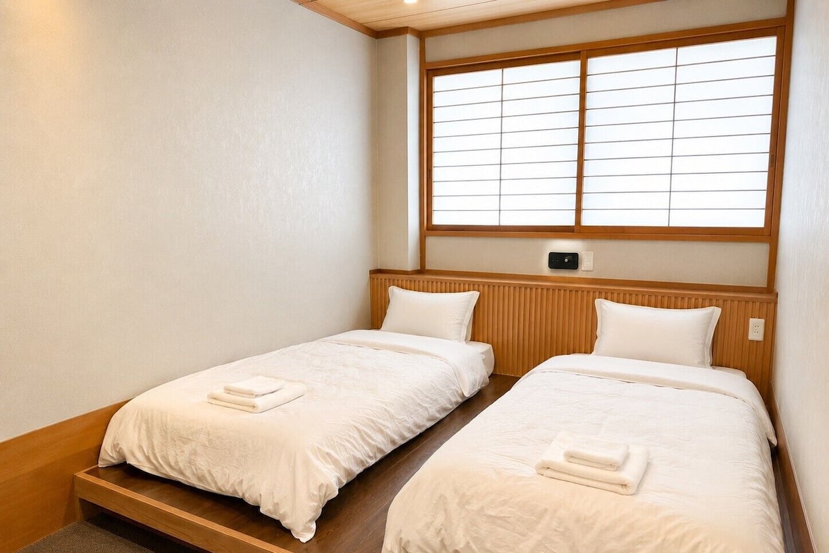 A room with two beds, wooden headboards, a window with blinds, and a small control panel on the wall.