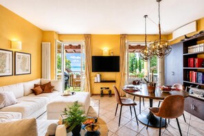 A modern living room with a sofa, a dining area with a table and chairs, a TV mounted on the wall, and a bookshelf. - Alice Suite Terrace, lake view & Park, Gardone Riviera, Italy (Gardone Riviera)