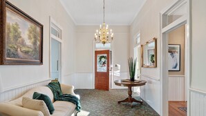 A hallway with a white ceiling, a chandelier, a wooden door with a wreath, a round wooden table, and a couch with a green throw blanket.