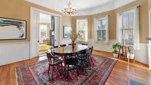 A dining room with a wooden table and chairs, a chandelier, and a large window with blinds.