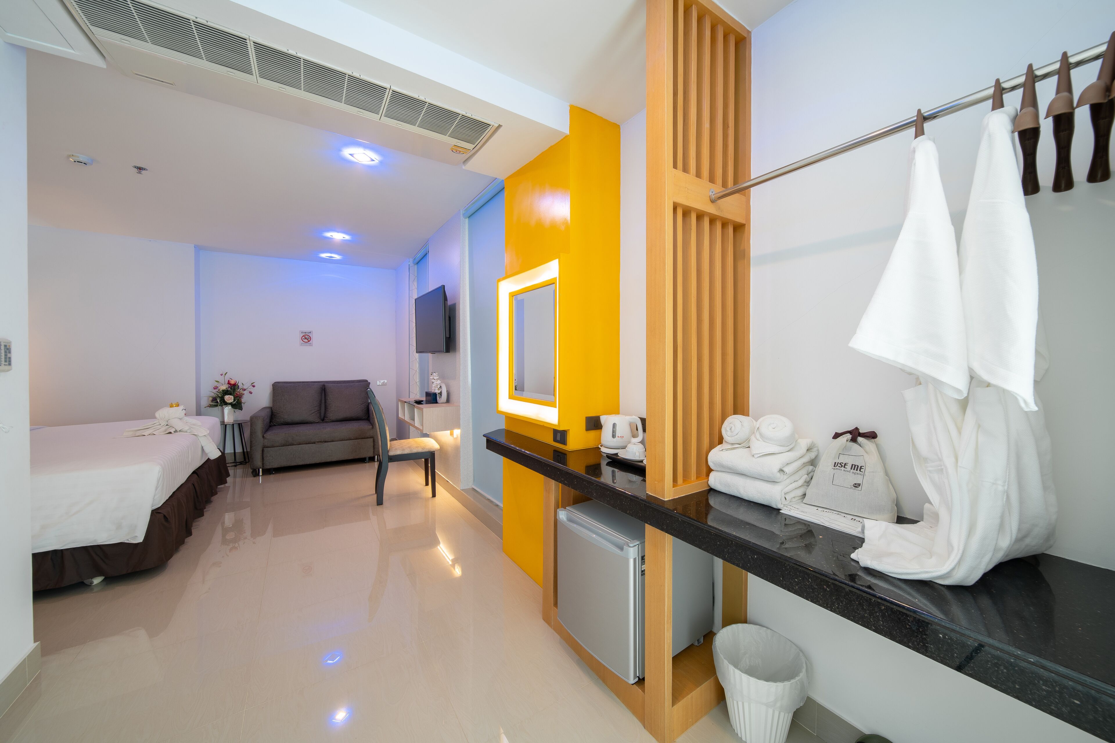 A hotel room with a bed, a sofa, a small table with a vase, a mirror, a towel rack, and a mini-fridge.