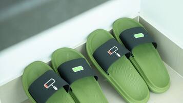A pair of green slippers with a black strap and a battery icon, placed on a light-colored floor next to a white wall.