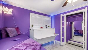 Un dormitorio con paredes moradas, un escritorio blanco, una cama con manta morada y un letrero de neón en la pared.