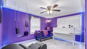 Una habitación con paredes moradas, ventilador de techo, cómoda blanca y cama con colchón negro.