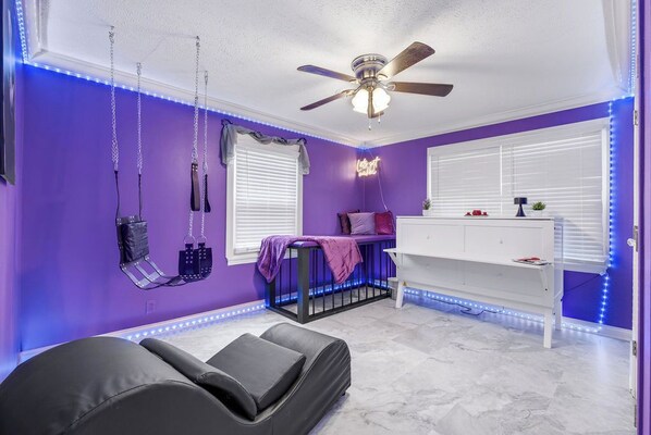 Una habitación con paredes moradas, ventilador de techo, cómoda blanca y cama con colchón negro.