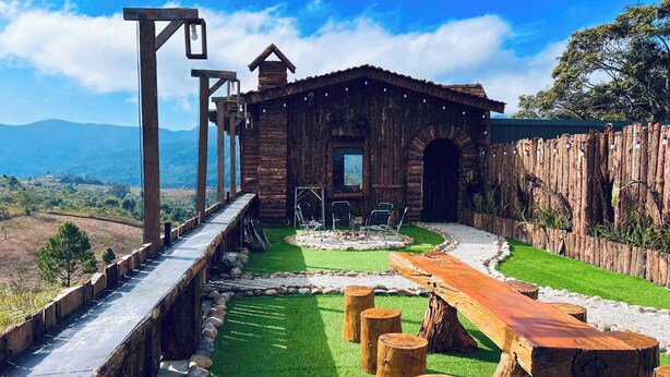 A rustic cabin with a stone pathway leading to a wooden deck, surrounded by a wooden fence and a view of distant mountains. - Gió Homestay – Măng Đen (Mang Den)