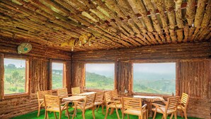 A rustic wooden room with a green artificial grass floor, wooden tables and chairs, and large windows offering a view of the countryside. - Gió Homestay – Măng Đen (Mang Den)