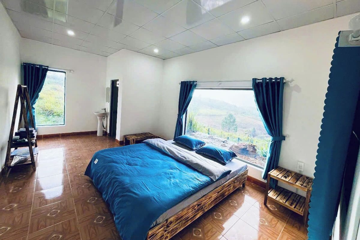 A bedroom with a large bed, a view of the outdoors, a wooden floor, and a window with curtains.