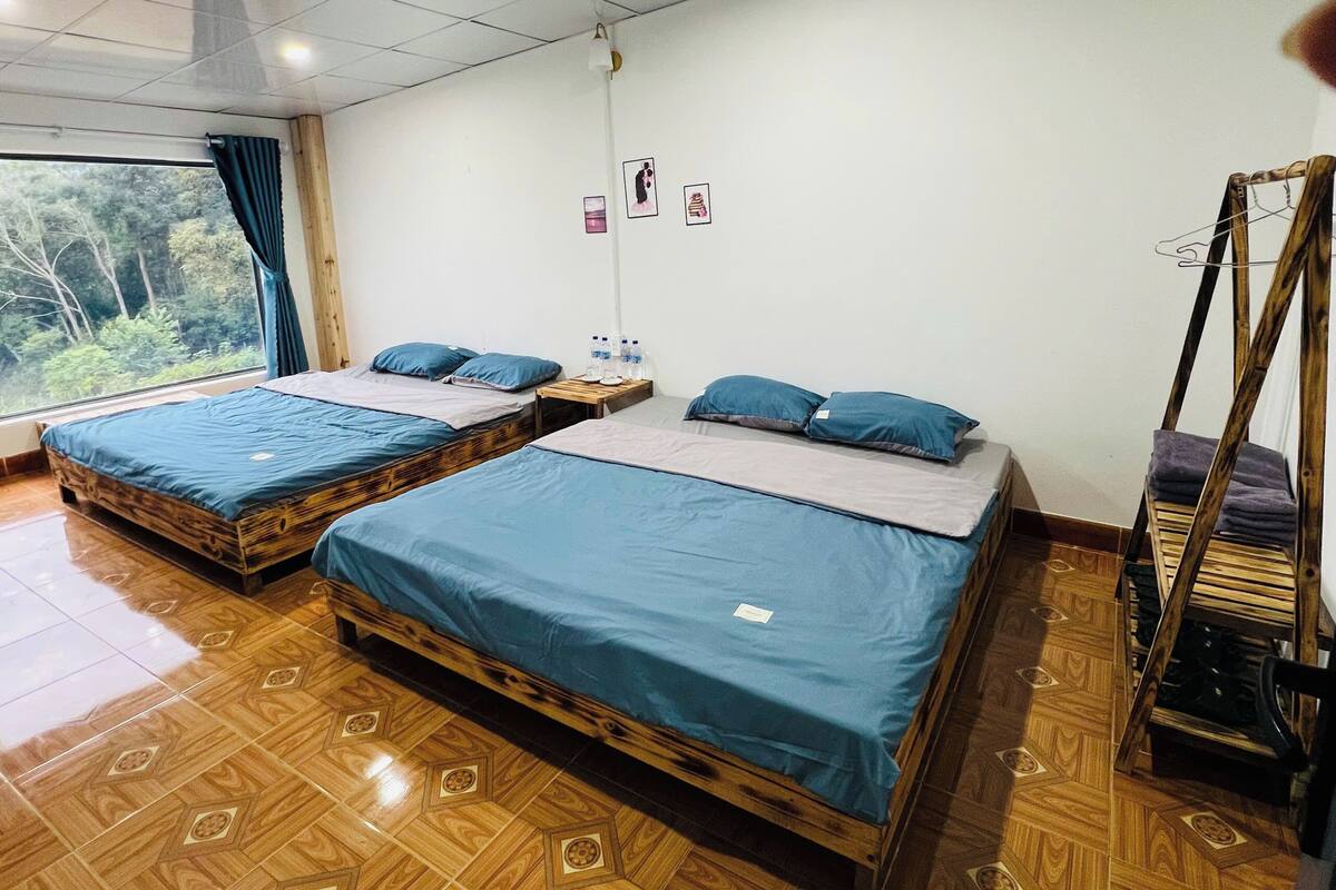 A room with two wooden beds, a wooden wardrobe, a window with curtains, and a wall-mounted light fixture.