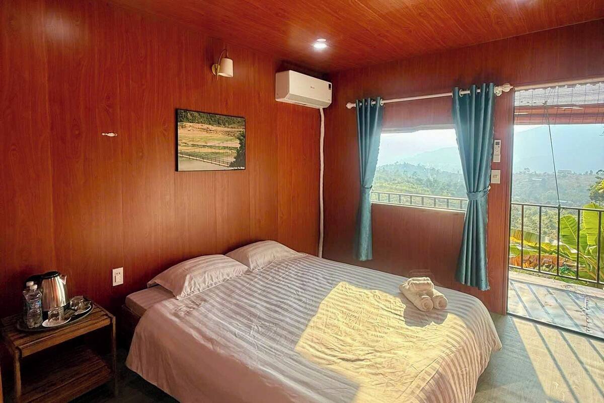 A wooden-paneled bedroom with a bed, a nightstand, an air conditioning unit, and a view of greenery through a balcony.