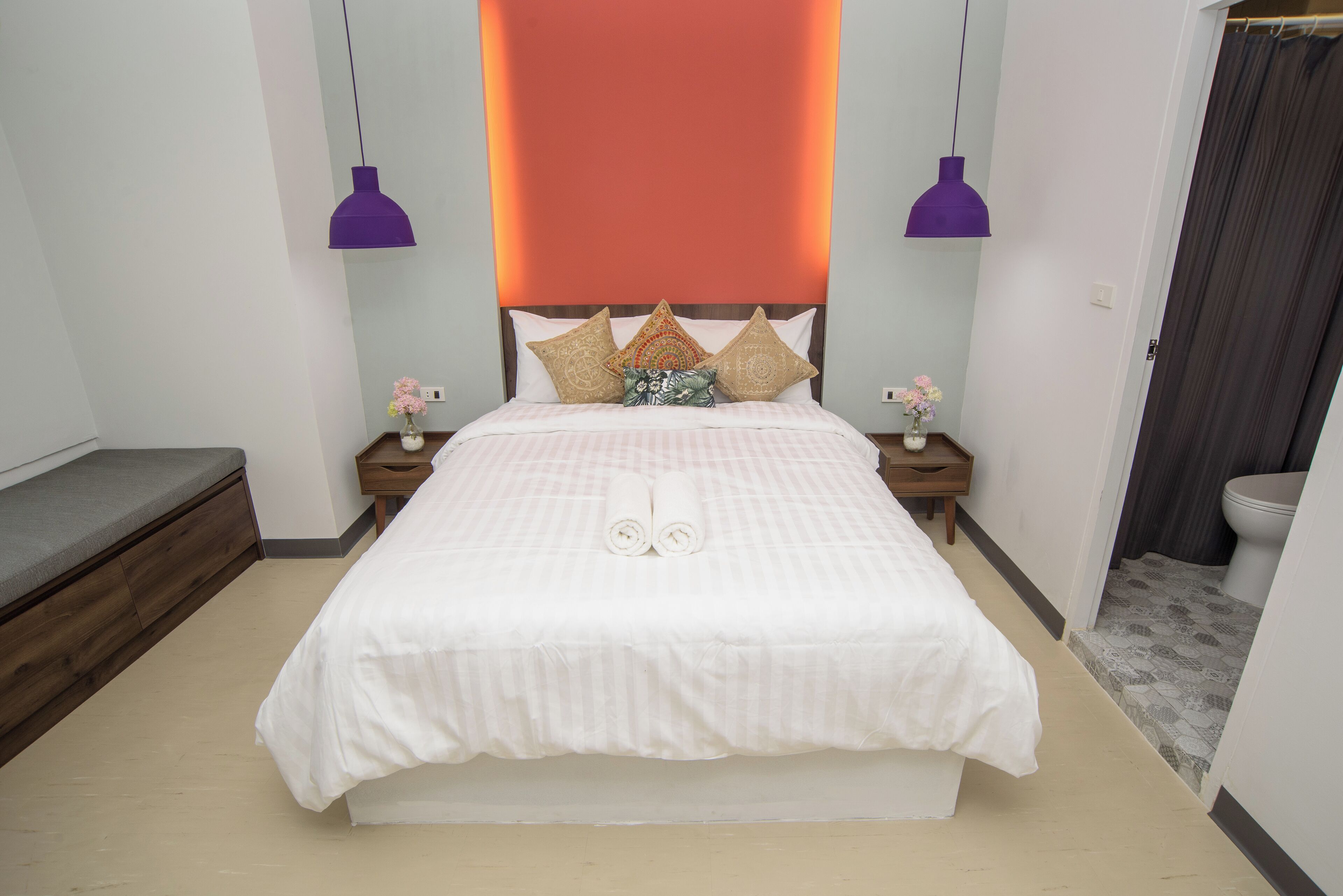 A neatly made bed with two rolled towels, a bedside table with a vase of flowers, and a nightstand with a lamp.