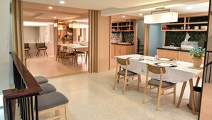 Un comedor con una mesa dispuesta para cuatro personas, rodeada de sillas. A la derecha hay una escalera y un área de cocina al fondo.