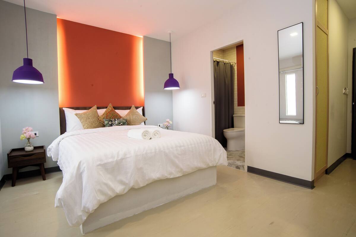 A modern bedroom with a large bed, two pendant lights, a nightstand with a vase, and a bathroom visible through an open door.