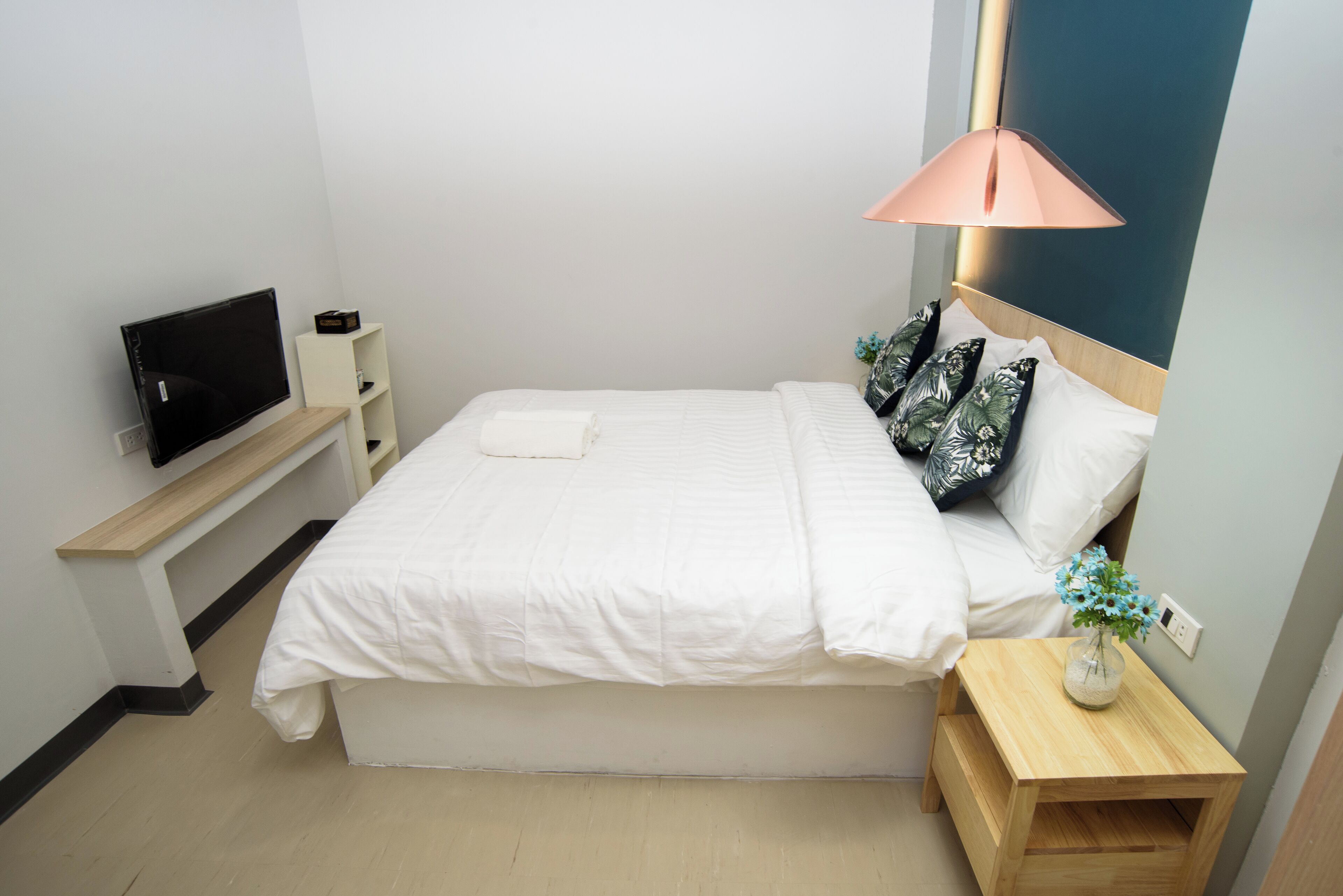 Una cama bien hecha con ropa de cama blanca, una mesita de noche con un florero y flores, un televisor sobre un mueble, y una lámpara fijada a la pared.