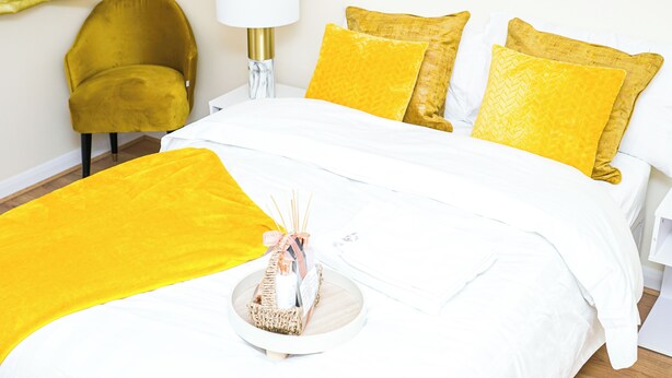 A neatly made bed with white bedding and multiple yellow pillows, a small bedside table with a lamp, and a mustard-colored chair.