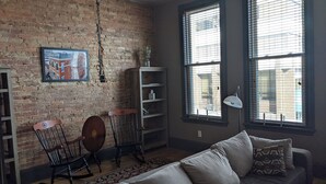 Een kamer met een bakstenen muur, een houten schommelstoel, een boekenkast en een raam met zonwering.