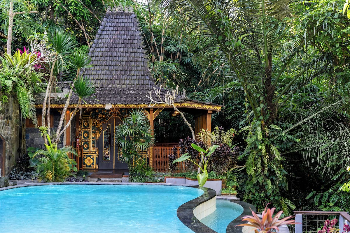 A pool with a tropical setting, surrounded by lush greenery and a small building with a tiled roof.