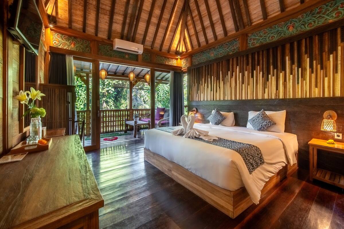 A spacious bedroom with a large bed, wooden flooring, and a balcony with a view of greenery.