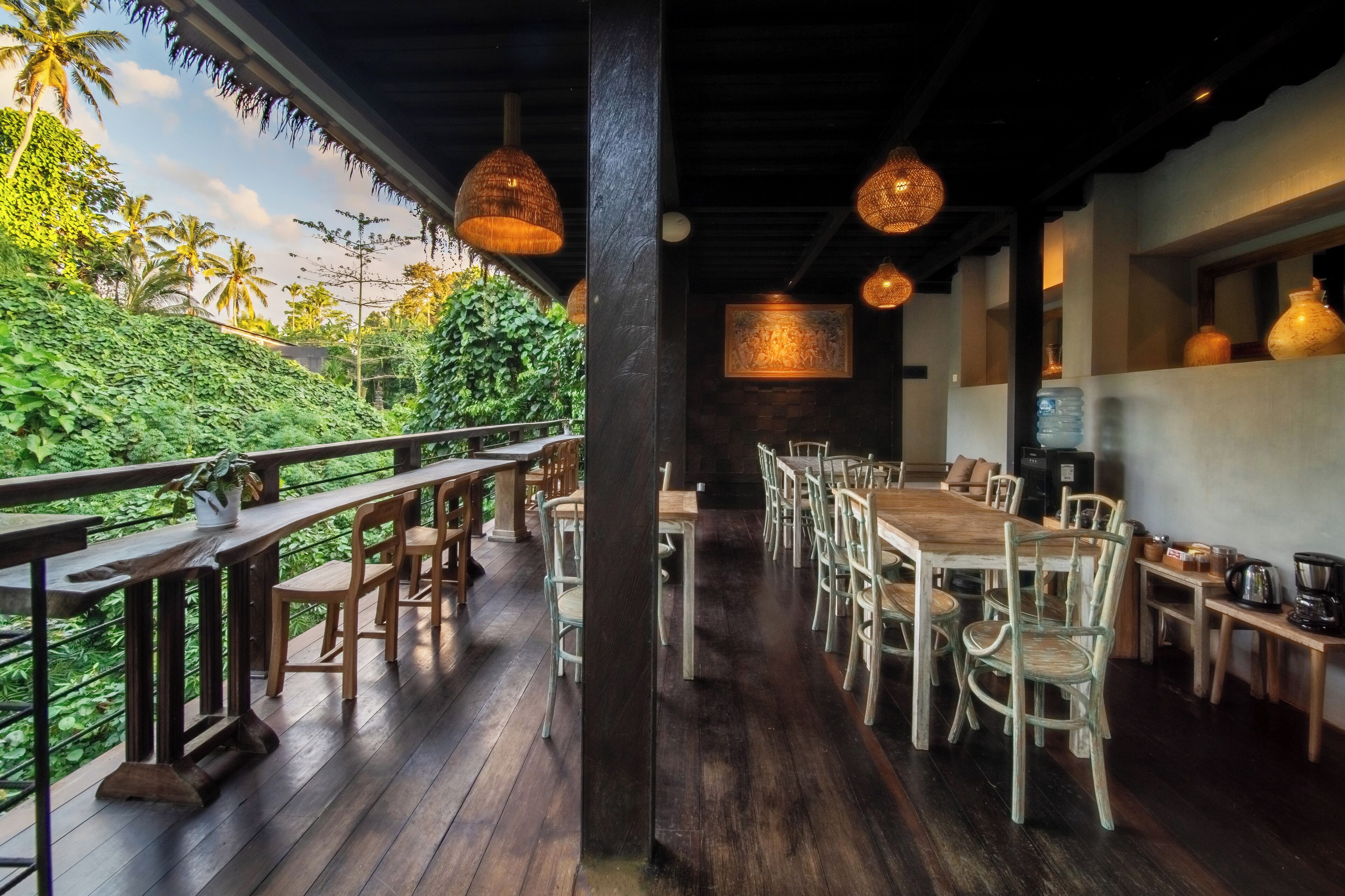 An outdoor dining area with wooden tables and chairs, surrounded by lush greenery and palm trees.