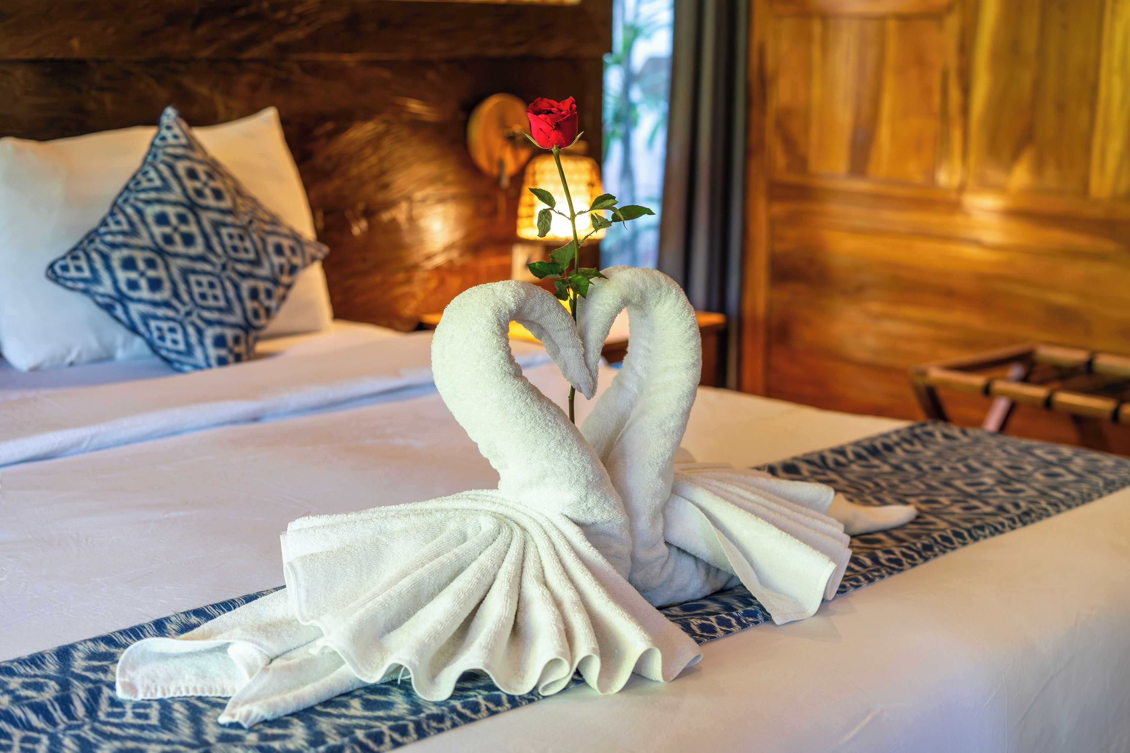 Un lit bien fait, agrémenté d’une serviette décorative disposée en forme de deux cygnes tenant une rose, d’un coussin à motifs et d’une tête de lit en bois.