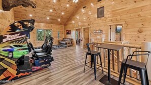 A wooden cabin interior with a moose head mounted on the wall, a racing game machine, a sofa, and a bar area.