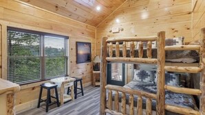 A wooden cabin interior with a bunk bed, a desk, two stools, a window with blinds, and a lamp.