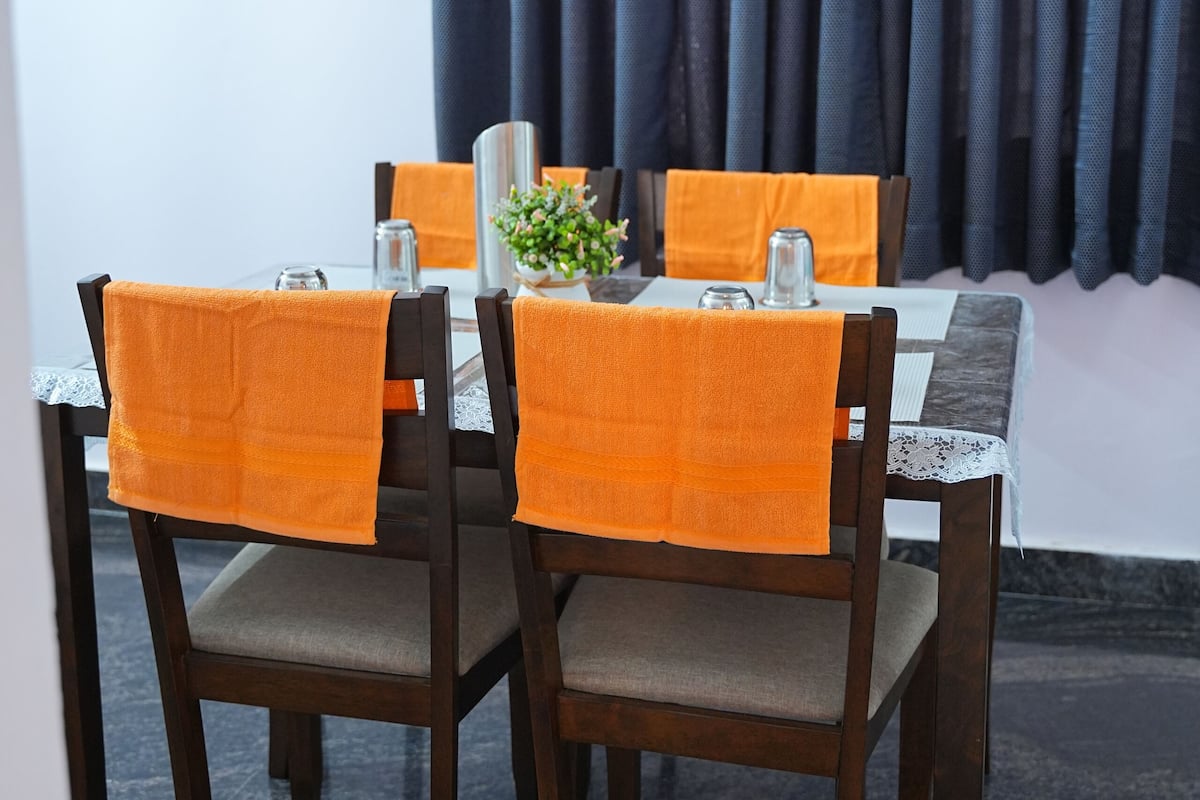 Una mesa de comedor dispuesta con cuatro sillas, cada una con una toalla naranja. La mesa está cubierta con un mantel blanco, y hay dos jarrones sobre ella.