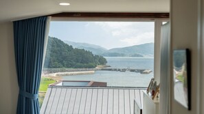A room with a large window overlooking a body of water, a dock, and a forested hillside. - That Day's Namhae Ocean View Pension (Namhae)