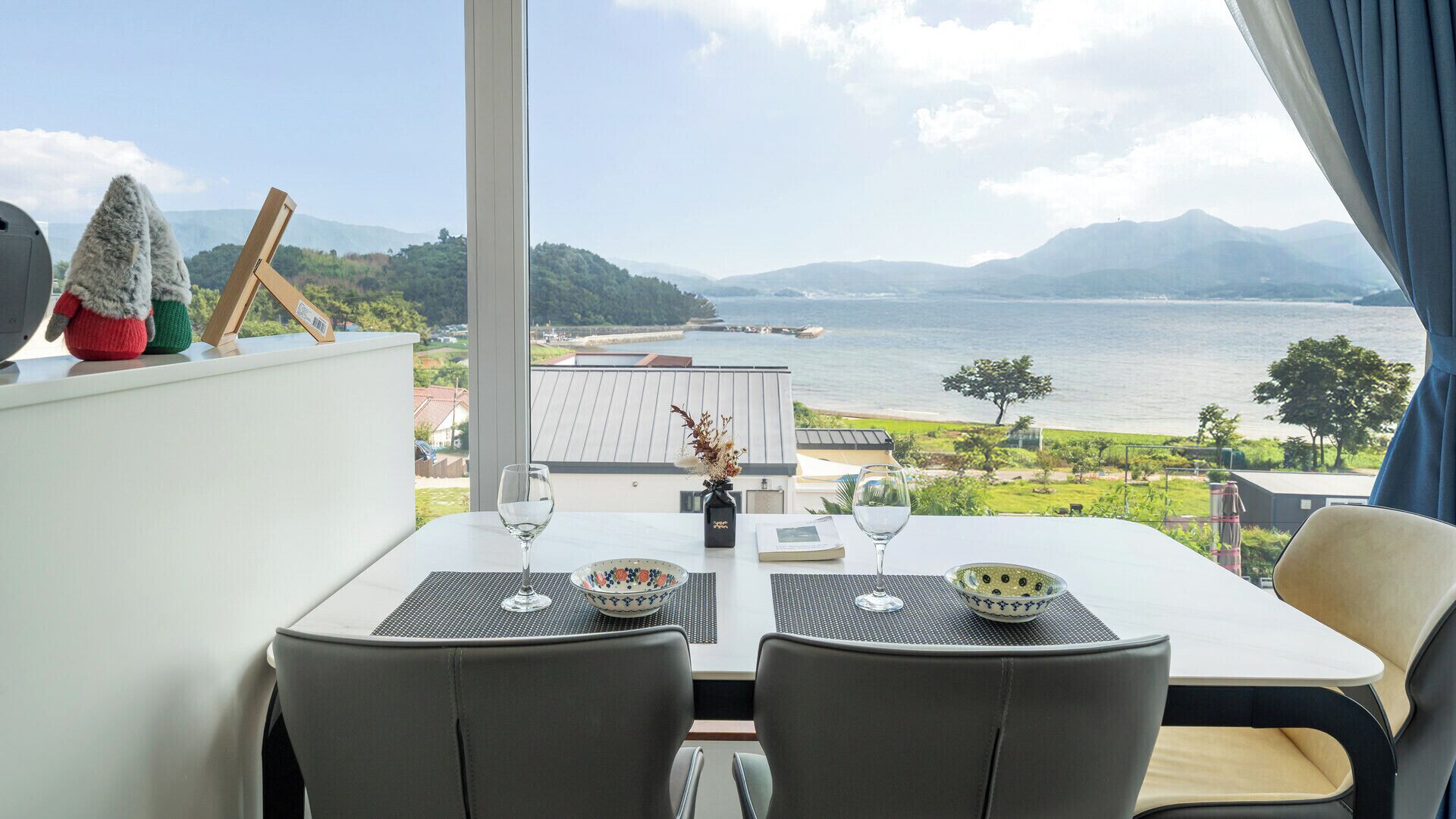 A dining table with two chairs, a vase, and a bowl on a balcony overlooking a body of water and hills.