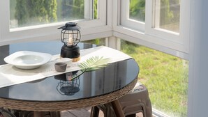 Una mesa redonda con tapa de vidrio negro, un plato blanco, una taza pequeña, una vela y una silla de mimbre.