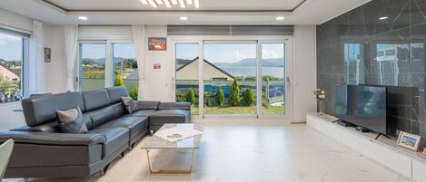 A modern living room with a sectional sofa, a flat-screen TV, and a large sliding glass door leading to a balcony with a view of the sea and mountains.