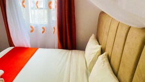 En säng med ett vitt och rött sängöverkast, en beige sänggavel och ett fönster med orangefärgade gardiner med lövmönster.