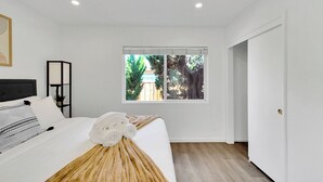 Ein Schlafzimmer mit Bett, einem Fenster mit Blick auf Bäume, einem Nachttisch und einer Tür.