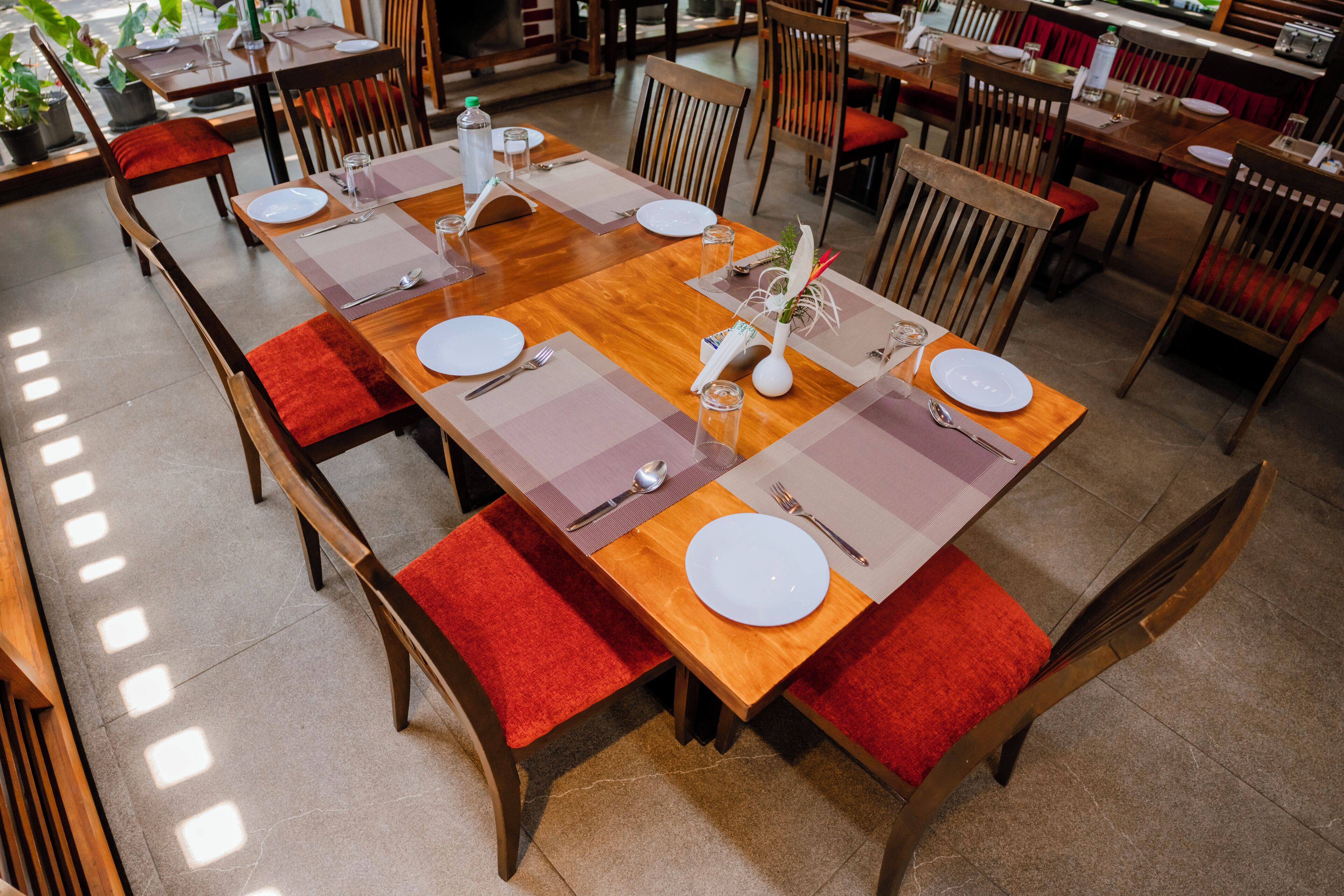A dining area with wooden tables, red cushioned chairs, and neatly set tables with plates, cutlery, and glasses.