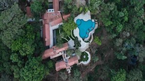 An aerial view of a property with a swimming pool, a large building with a red roof, and surrounding greenery.