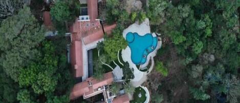 Vista aérea de una propiedad con piscina, un edificio grande de techo rojo y vegetación circundante.