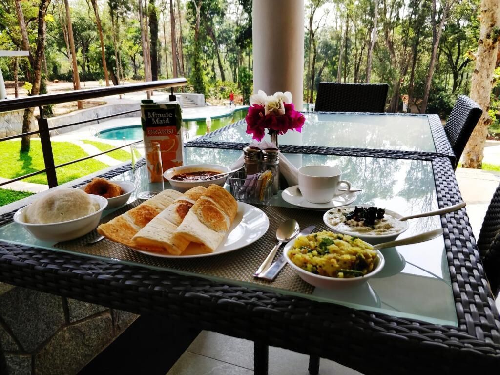 A table set with breakfast items, including bread, soup, and a cup, overlooking a pool and greenery.