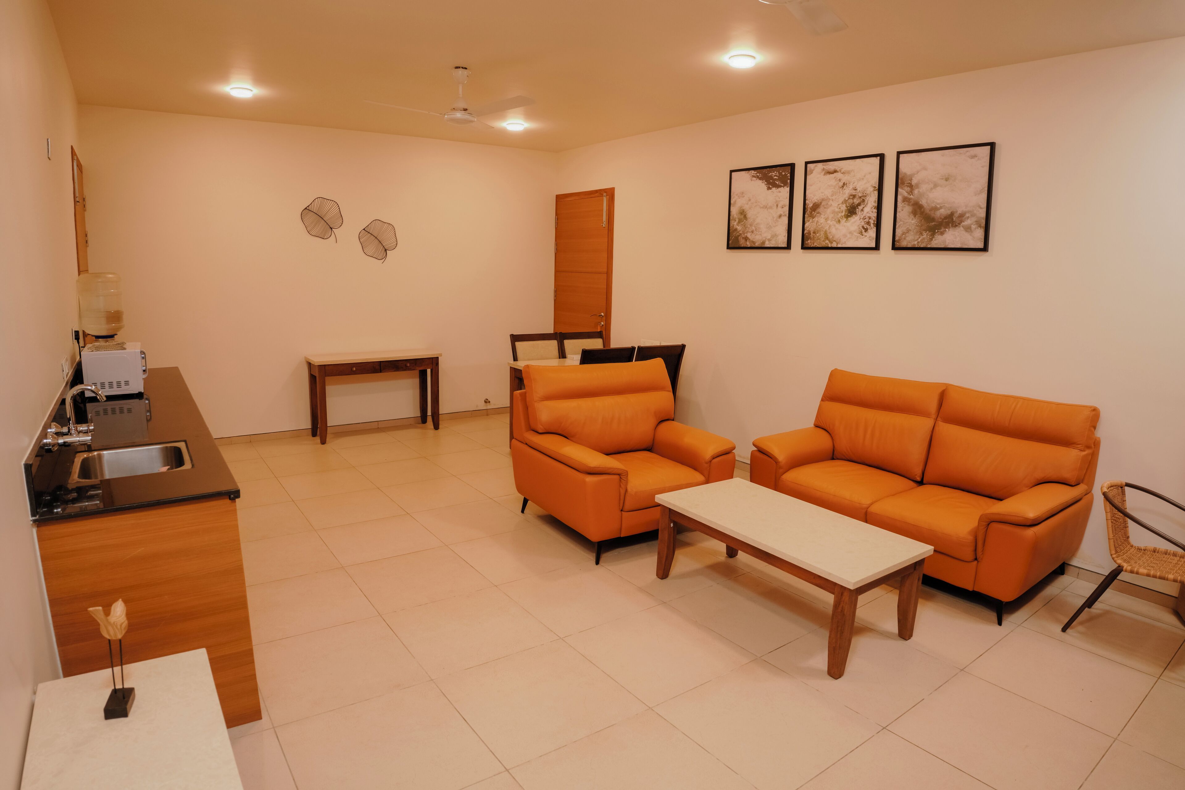 Ein modernes Wohnzimmer mit zwei orangefarbenen Sofas, einem Holztisch, einem kleinen Tisch mit einer Lampe und einer Küchenzeile mit Spüle und Wasserspender.