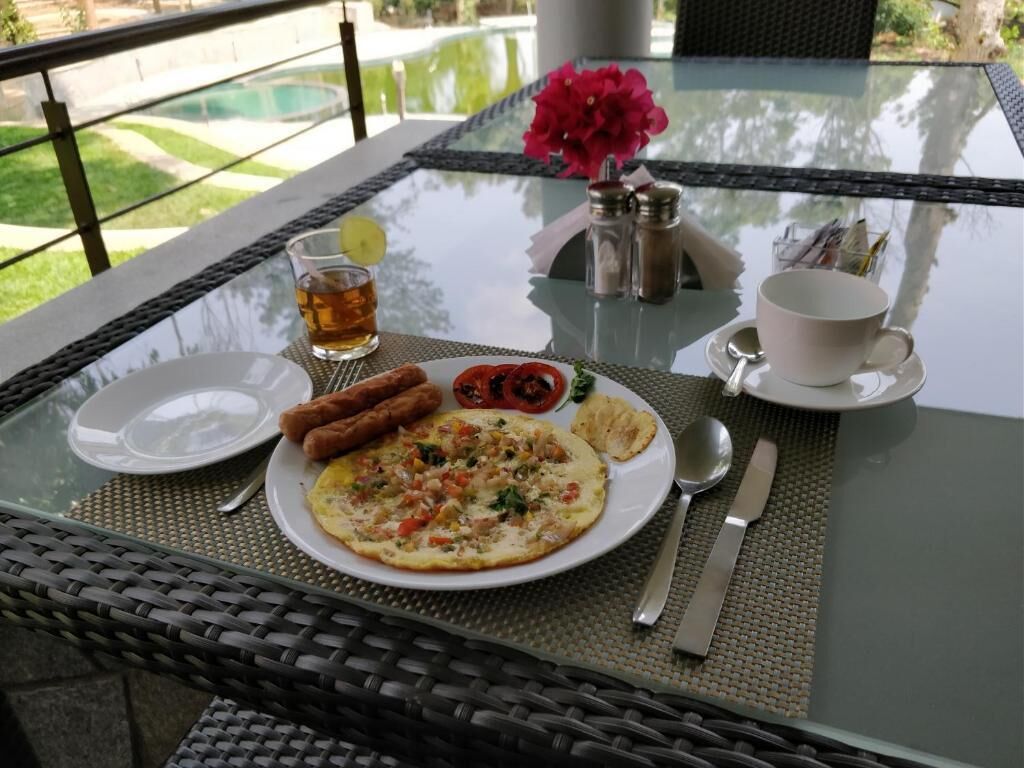 A breakfast table with a plate of food, a glass of tea, and a flower vase.