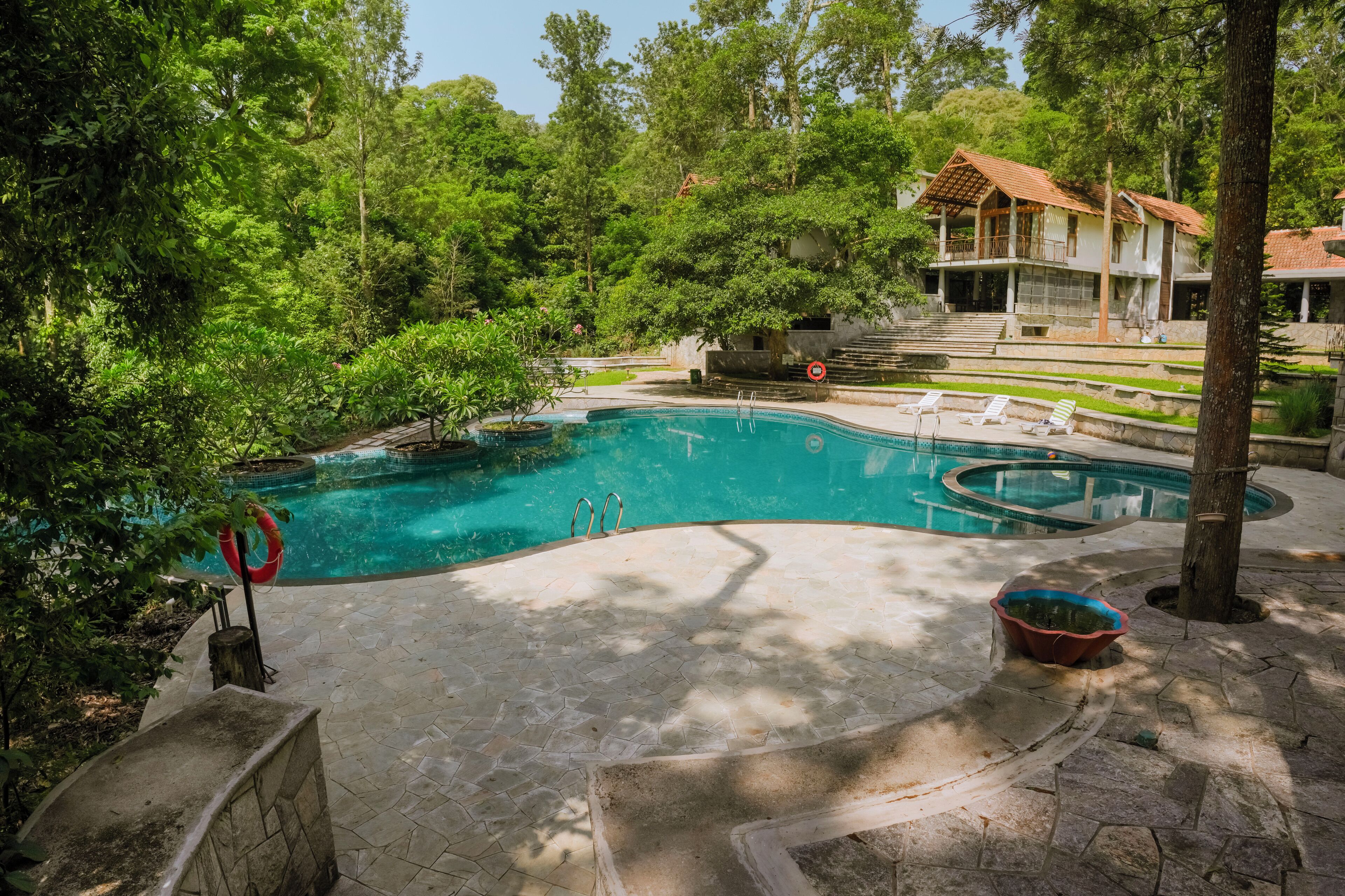 Piscina com deck de pedra, rodeada por vegetação exuberante e uma casa ao fundo.
