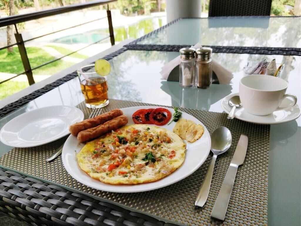 A breakfast setting with a plate of food, a cup, and a glass of tea on a glass table with a view of greenery.