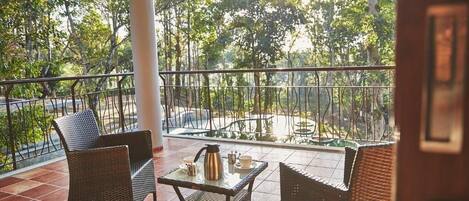 A balcony with wicker furniture and a table set with a teapot and cups, overlooking a garden with trees.