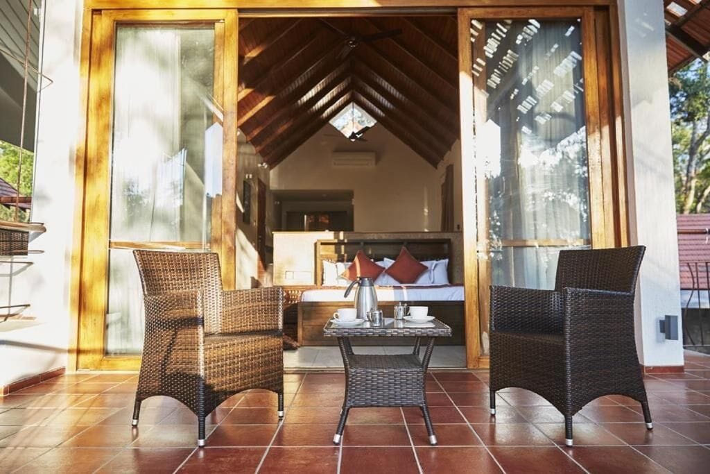 A room with a bed, a table with a kettle, and two wicker chairs.